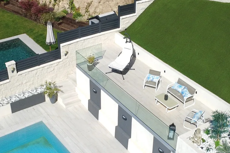 Vista aérea de una terraza elevada con barandilla de cristal sin montantes, tumbona y muebles de jardín, junto a la piscina