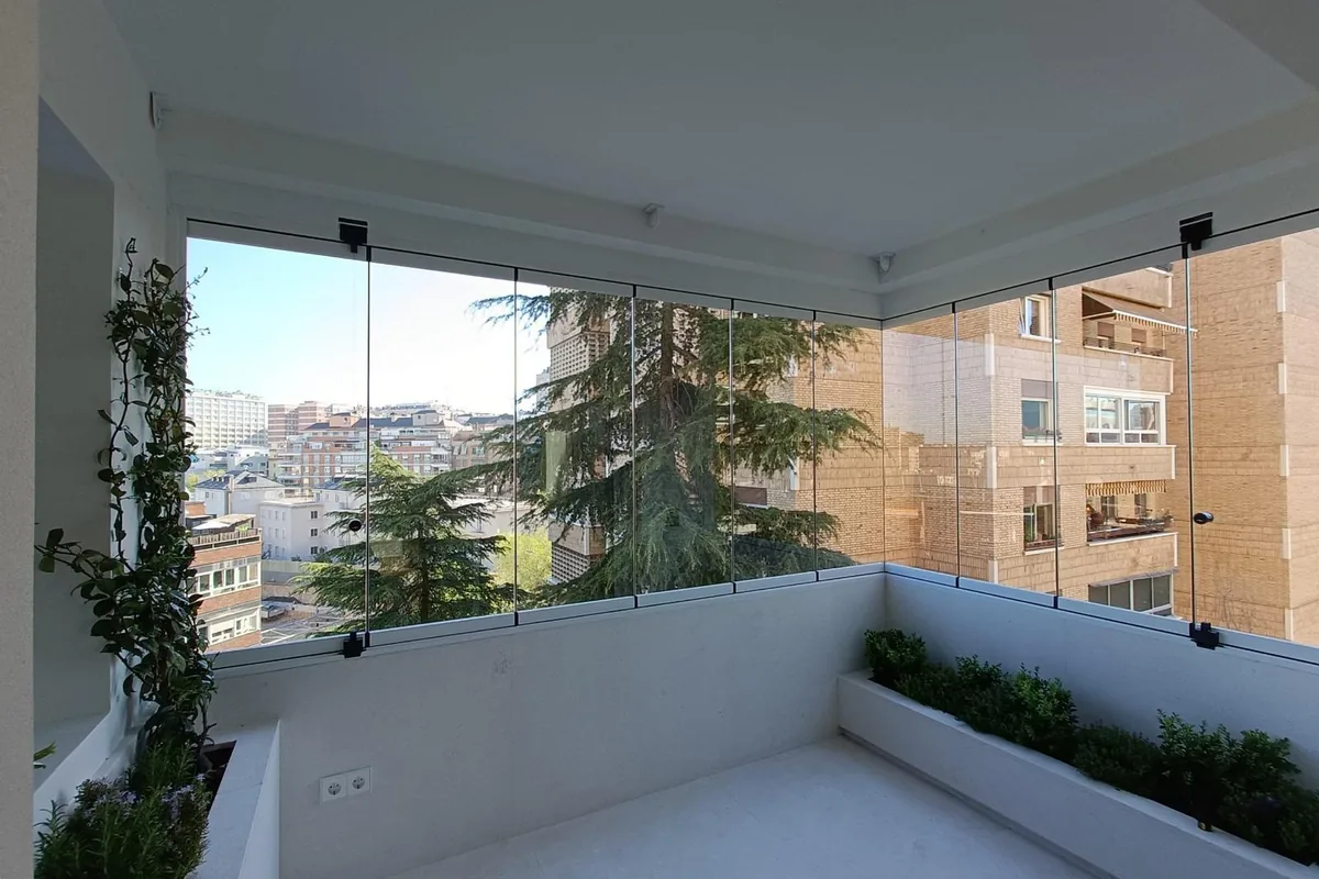 Cerramiento de cristal sin marco en esquina de balcón con plantas y vistas a la ciudad