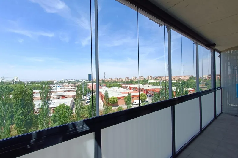 Cerramiento de cristal en esquina de ático con panel inferior translúcido y vistas a naves industriales