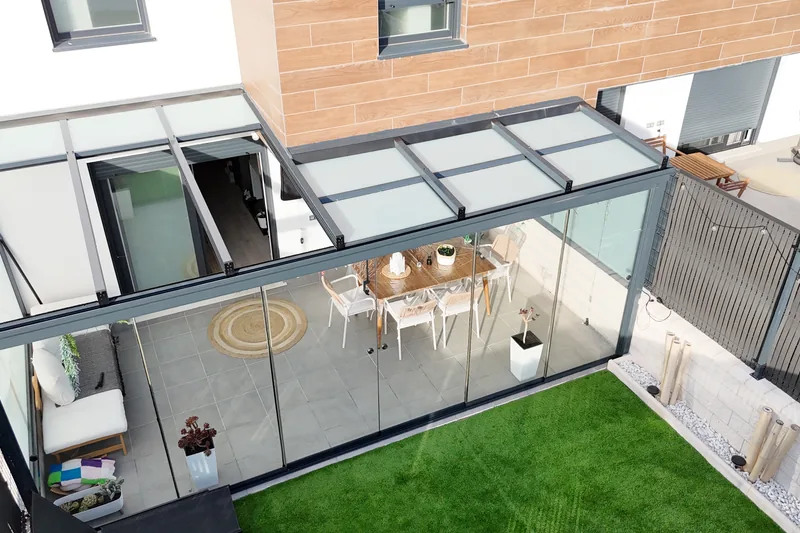 Vista aerea de un techo de cristal móvil sobre el patio de una vivienda, con los laterales cerrados con cristal, comedor exterior y césped