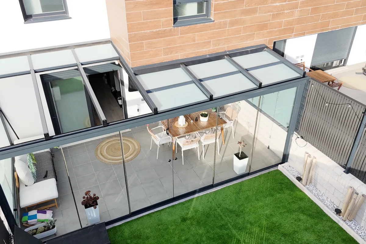 Vista aerea de un techo de cristal móvil sobre el patio de una vivienda, con los laterales cerrados con cristal, comedor exterior y césped
