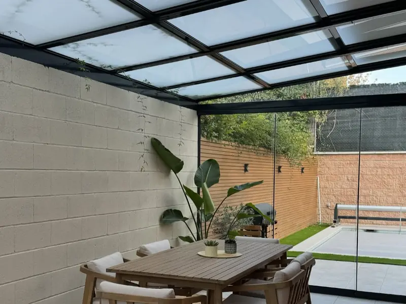 Comedor exterior bajo un techo de cristal, cerrado con una cortina de cristal corredera, junto a la piscina en Madrid