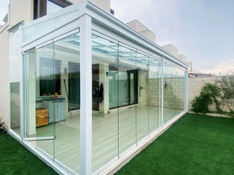 Porche cerrado con cortinas de cristal abatibles y techo de cristal, con perfilería lacada en blanco, en una vivienda de Madrid