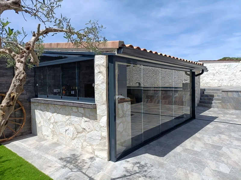 Cerramiento de cristal en porche con barra de piedra y vistas a jardin con olivo