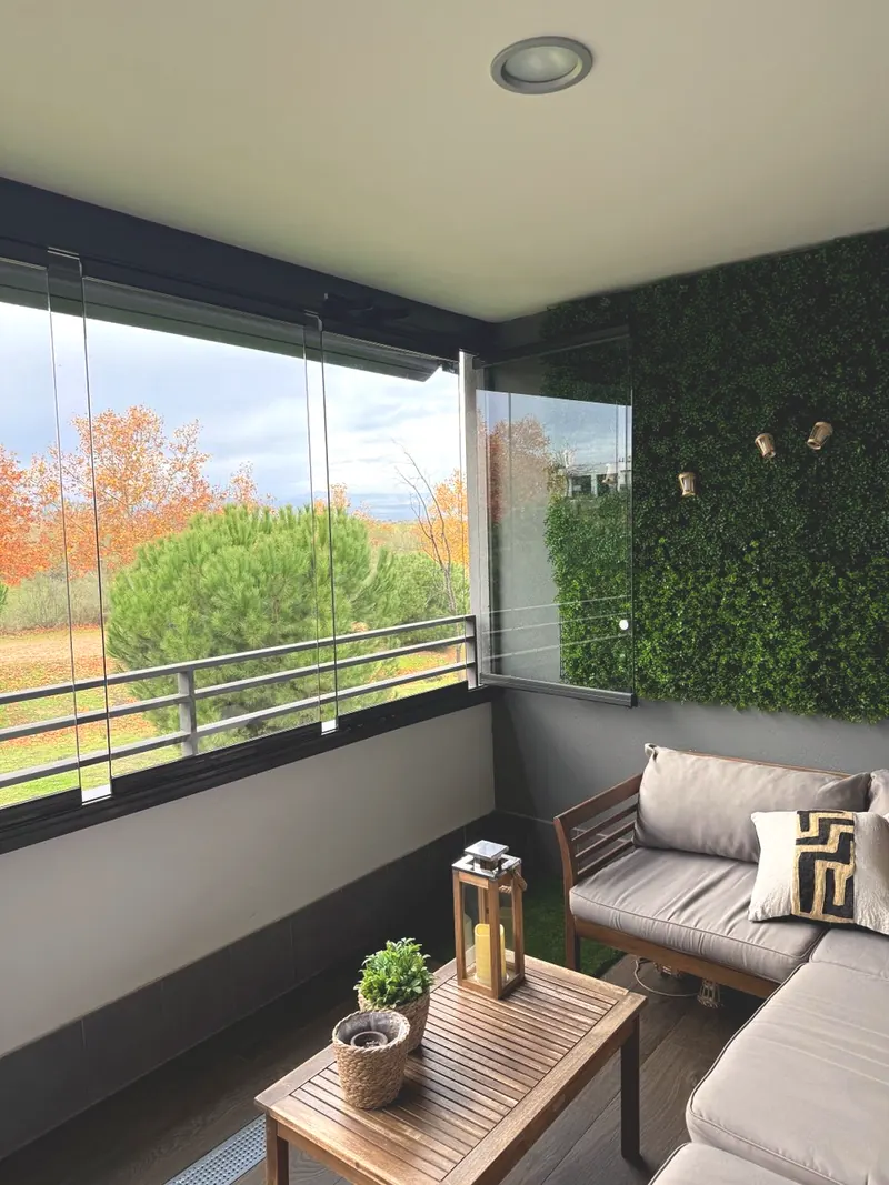 Terraza cerrada con cortinas de cristal, muro vegetal y sofá, con vistas al arbolado en otoño