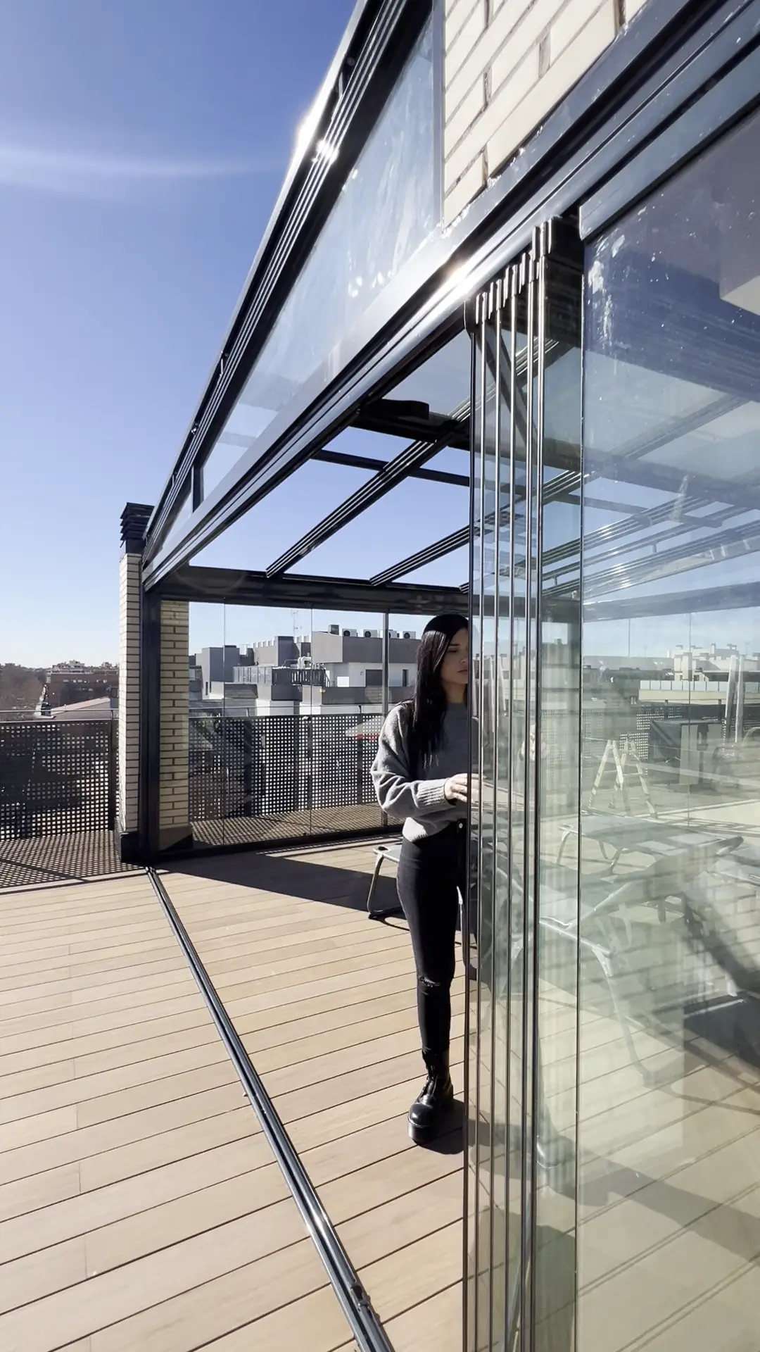 Mujer abriendo hoja a hoja una cortina de cristal abatible en la terraza de un ático de Madrid