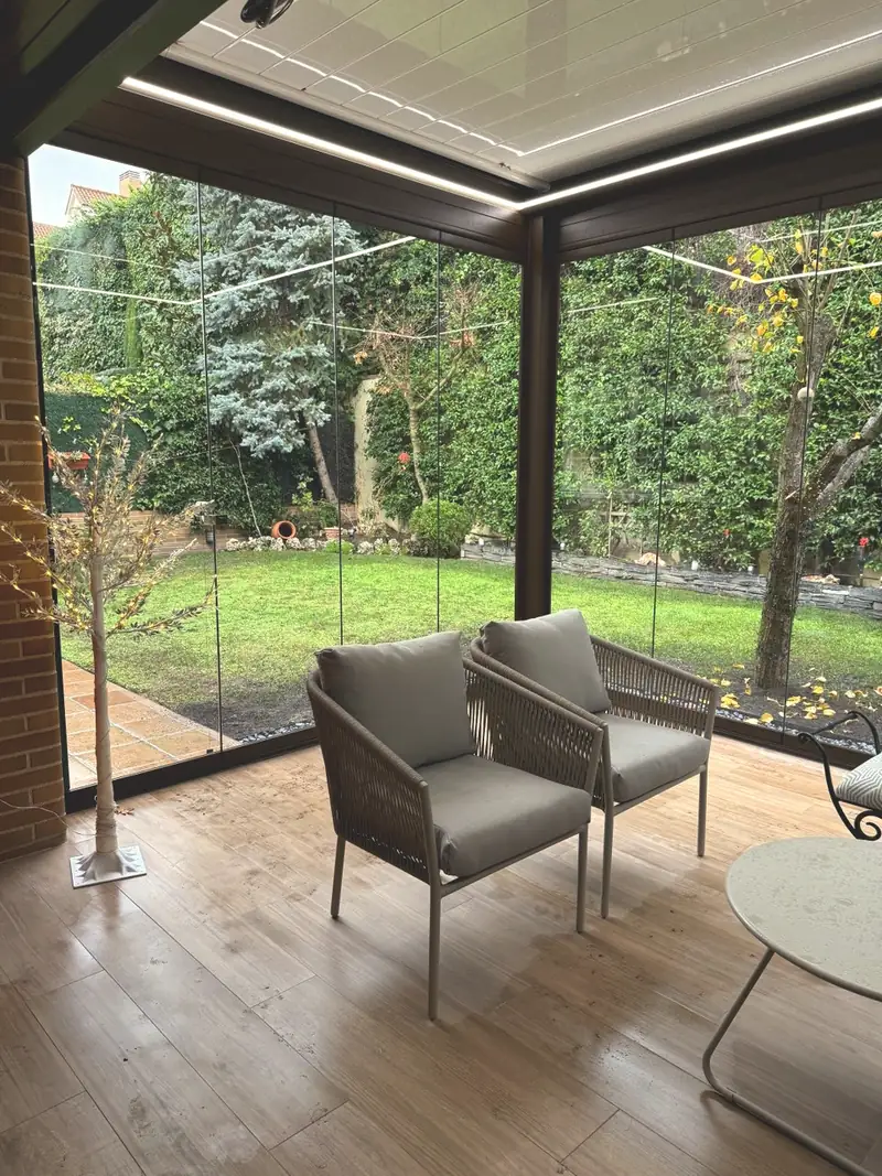 Interior de una pérgola bioclimática orientable con las lamas cerradas y luz LED integrada, con el jardín al otro lado del cristal