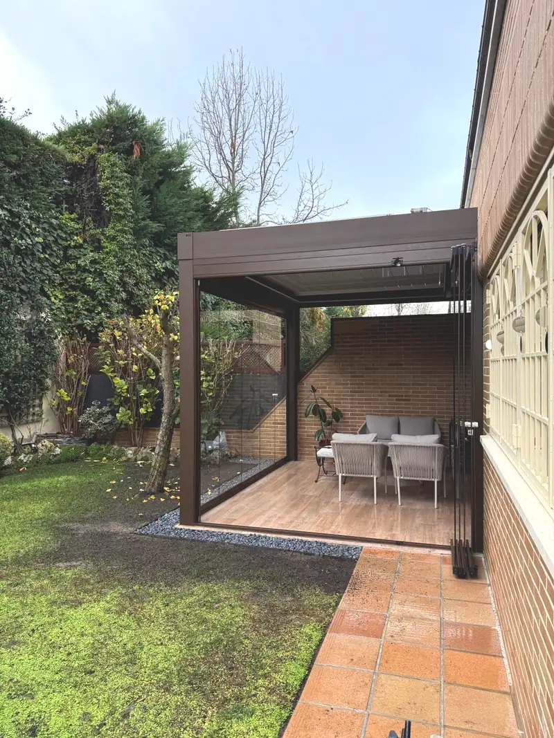 Pérgola bioclimática orientable con los laterales cerrados con cristal, vista desde el jardín de una vivienda de Madrid