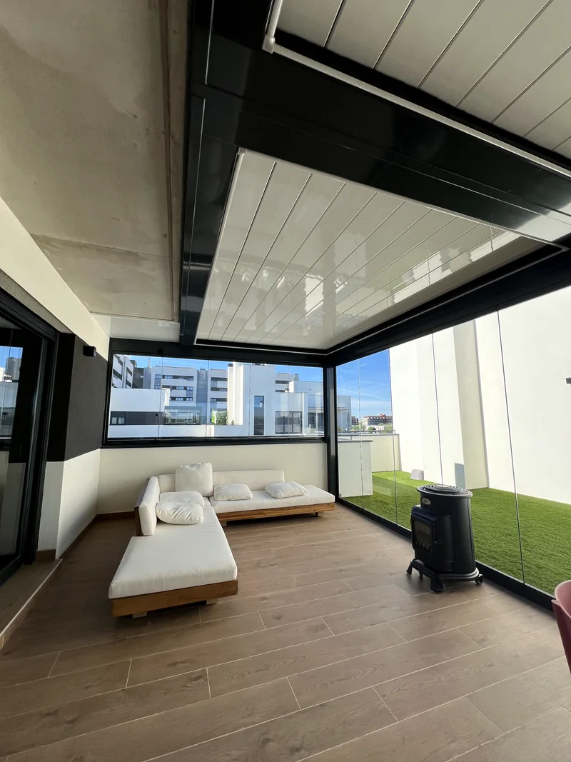 Salon exterior con pergola bioclimatica, sofa blanco, estufa y cerramiento de cristal sobre cesped artificial